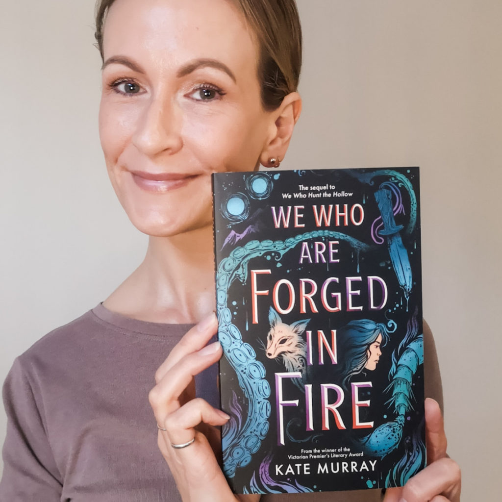 A smiling white woman with her hair pulled back holds up a book: We Who Are Forged In Fire